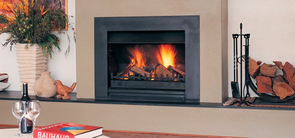 A Jetmaster Universal Insert built into a wall with stacked logs and a flower vase beside it, one of the best fireplaces for autumn warmth