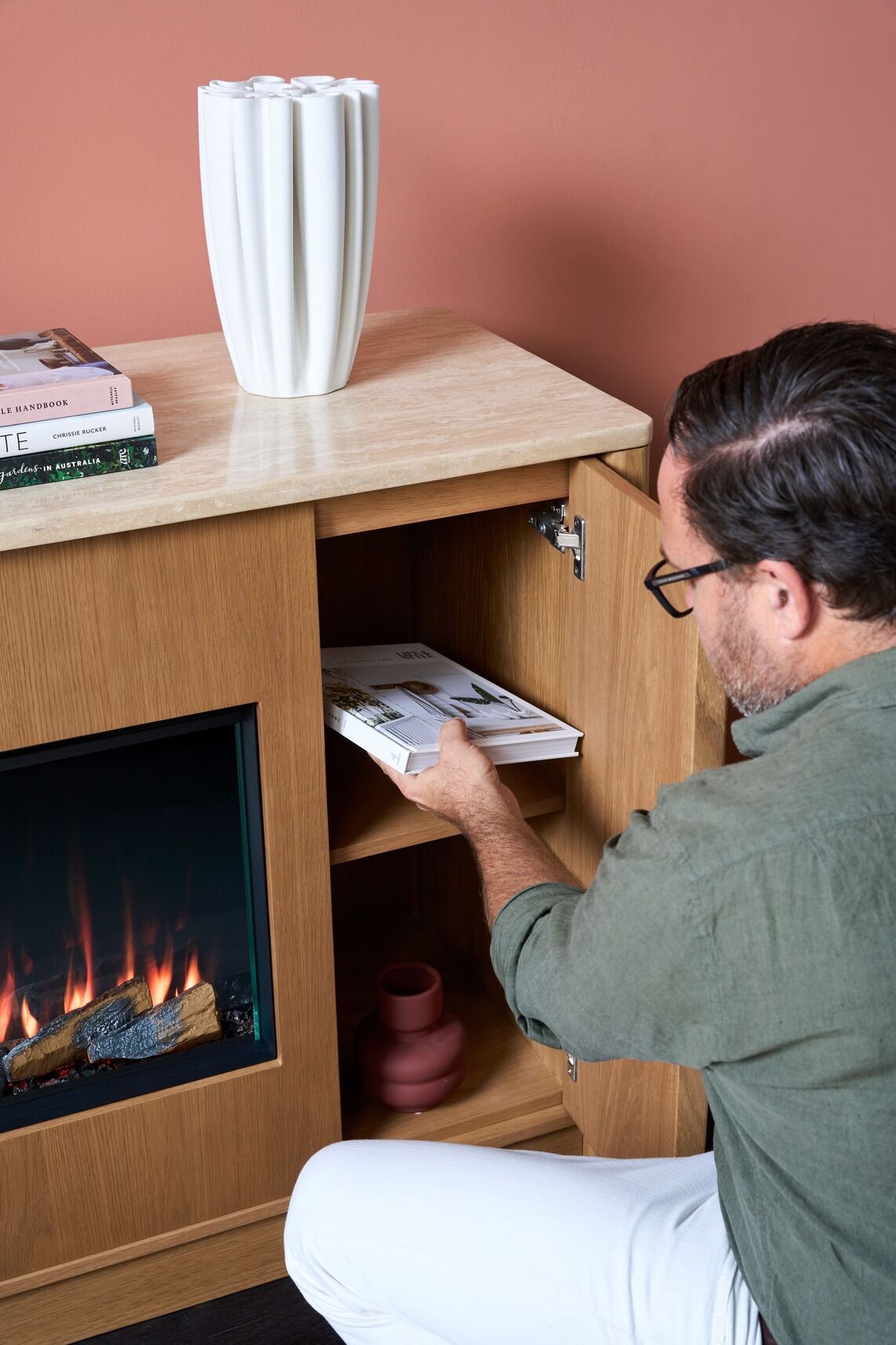 A man places a book inside a wooden cabinet beside the Ember Arc Grange 180 electric fireplace in a cosy indoor setting