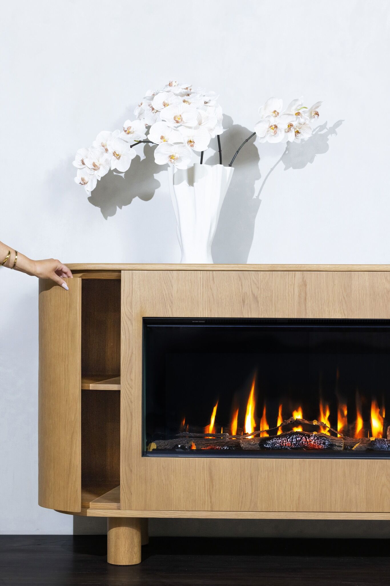 A person stands beside an electric fireplace, featuring a vase of flowers, with the EmberArc Ashton 180 prominently displayed